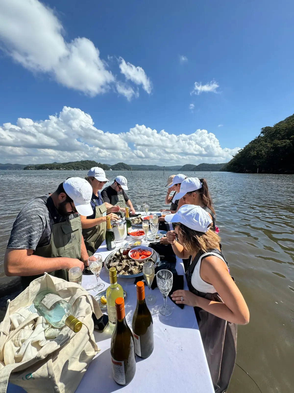 Guests in waders and Ahoy caps shucking oysters at a table set in the shallows of the Hawkesbury River