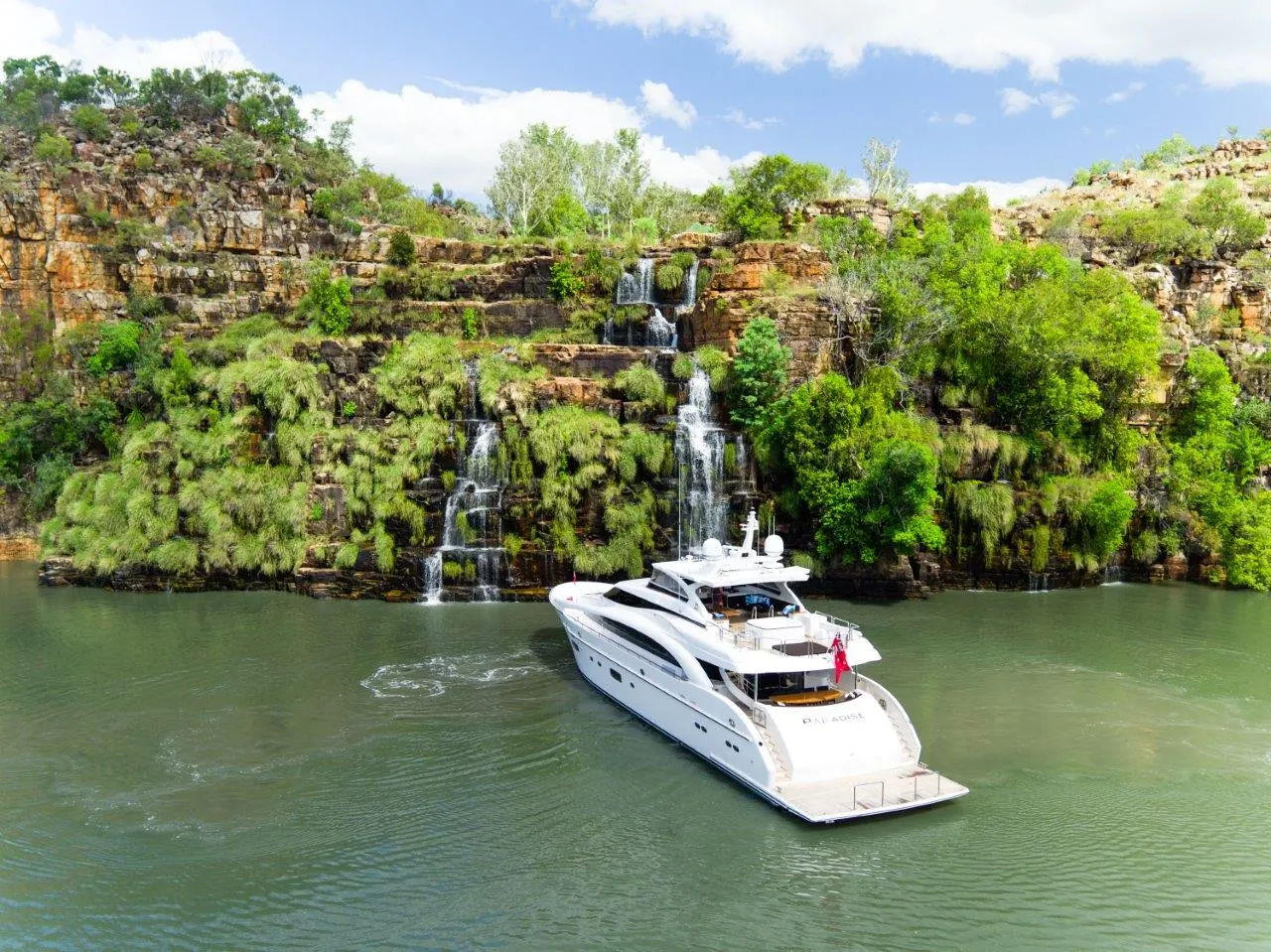 A motor yacht at anchor below waterfalls on a red sandstone cliff in the Kimberley