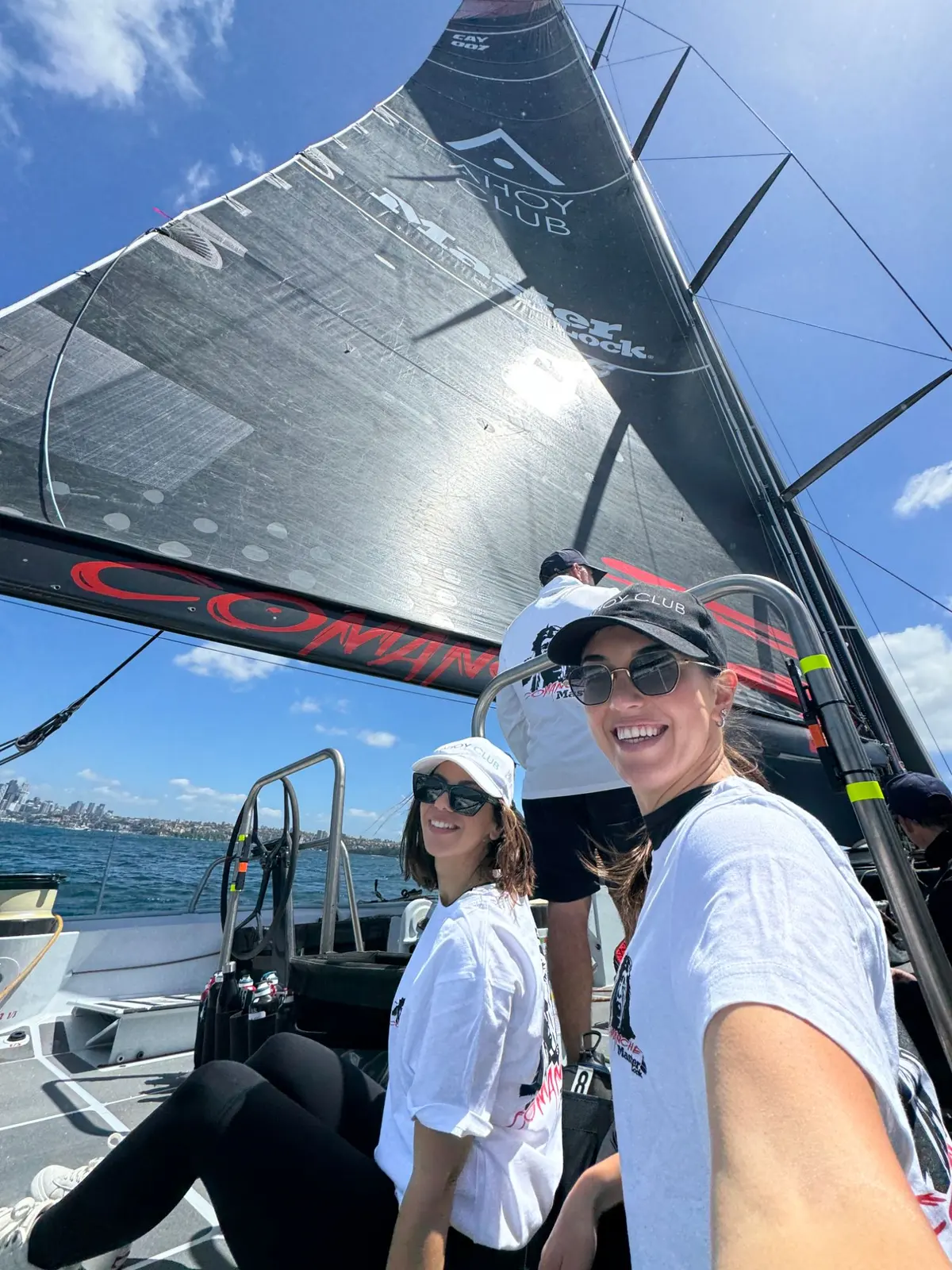 Two of the Ahoy Club team on the deck of Comanche under the Ahoy Club and Master Lock sail, Sydney Harbour behind