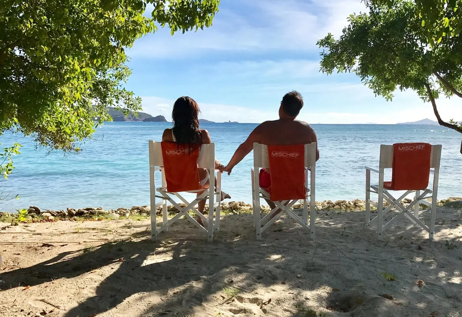 Two of the group in MISCHIEF deck chairs on the sand, looking out to the islands