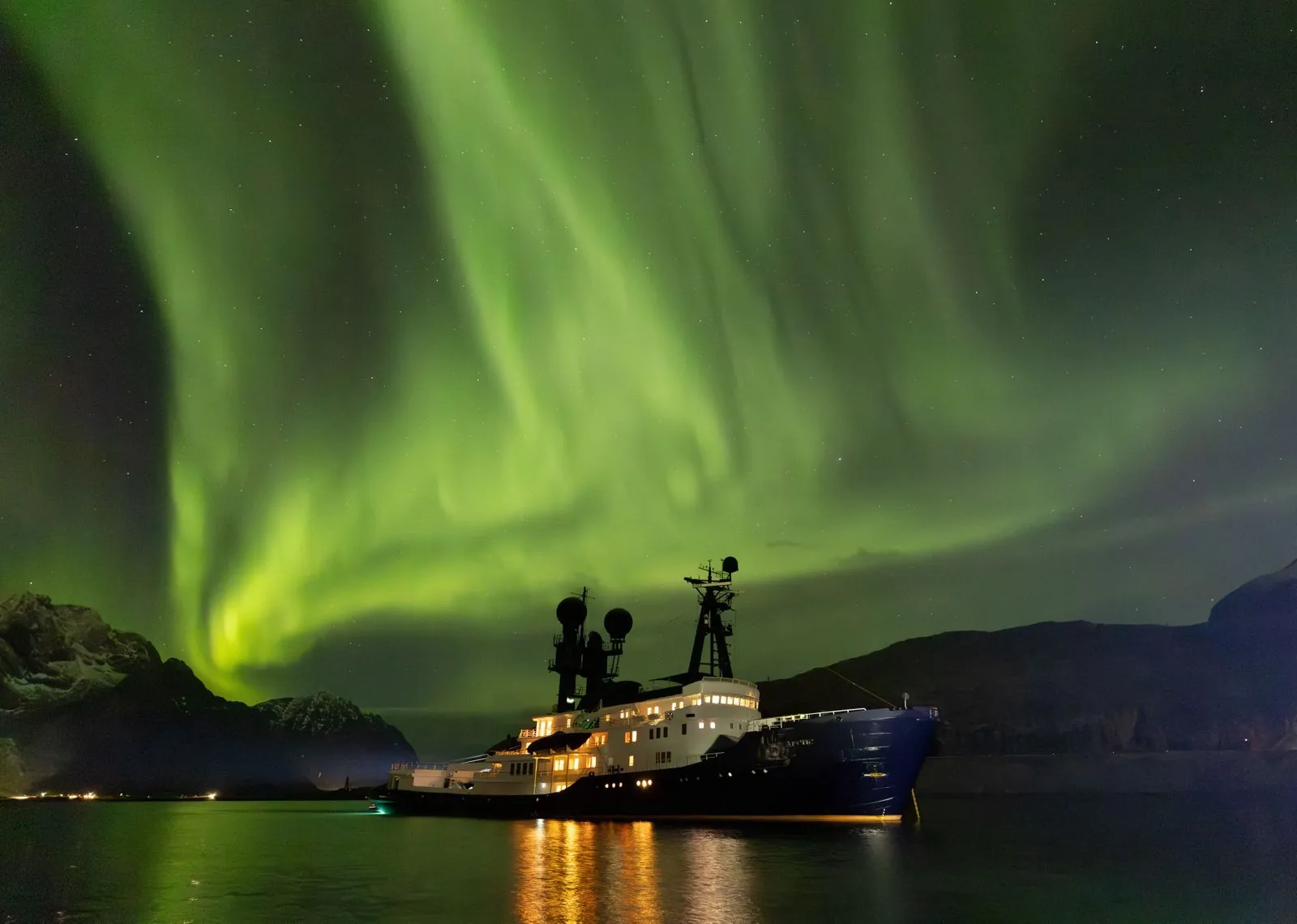 ARCTIC at anchor in a Norwegian fjord under the northern lights, her decks lit up and reflected in the water