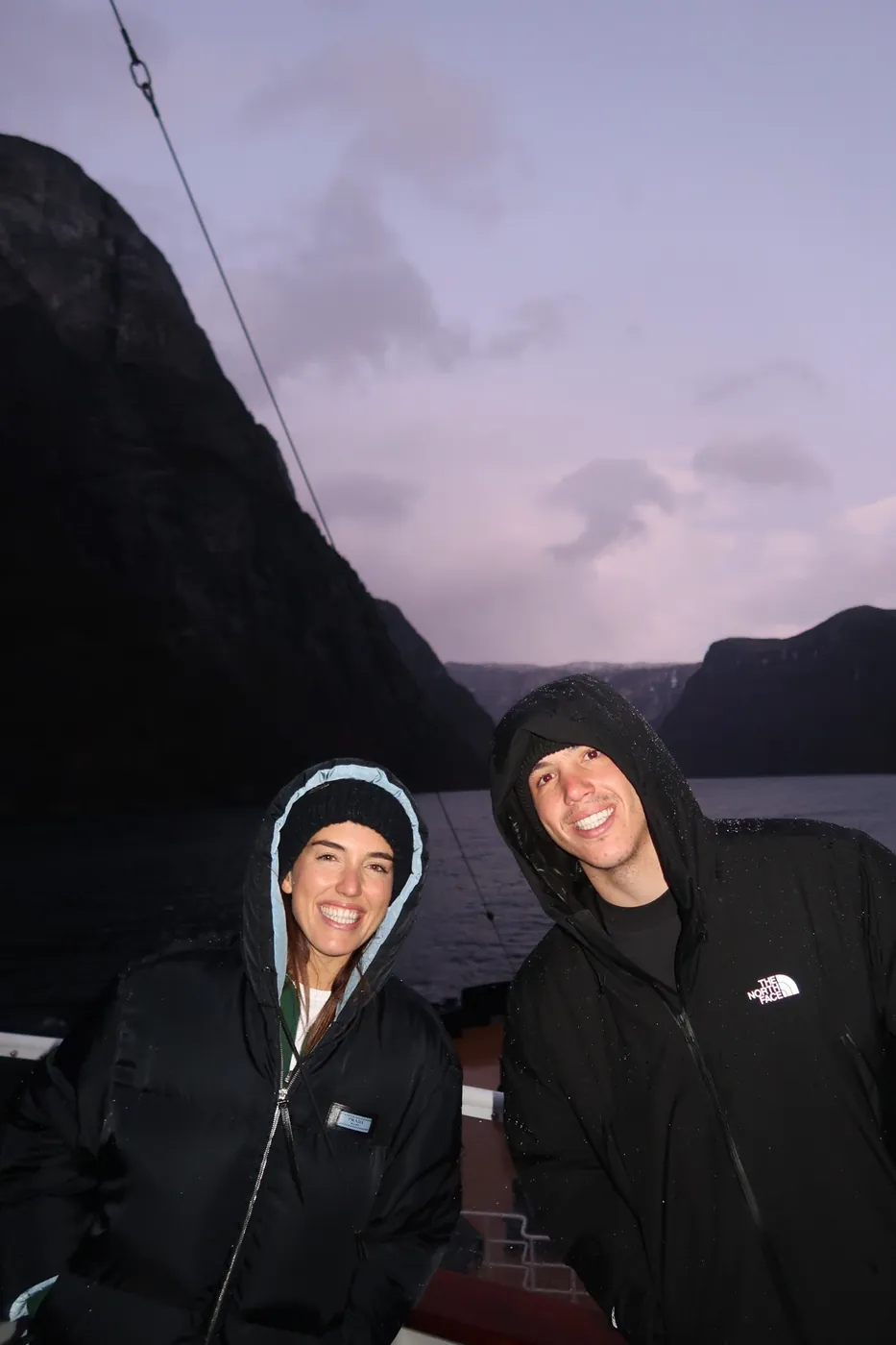 Ellie and a guest on deck at dusk, the fjord walls rising behind them under a lilac sky