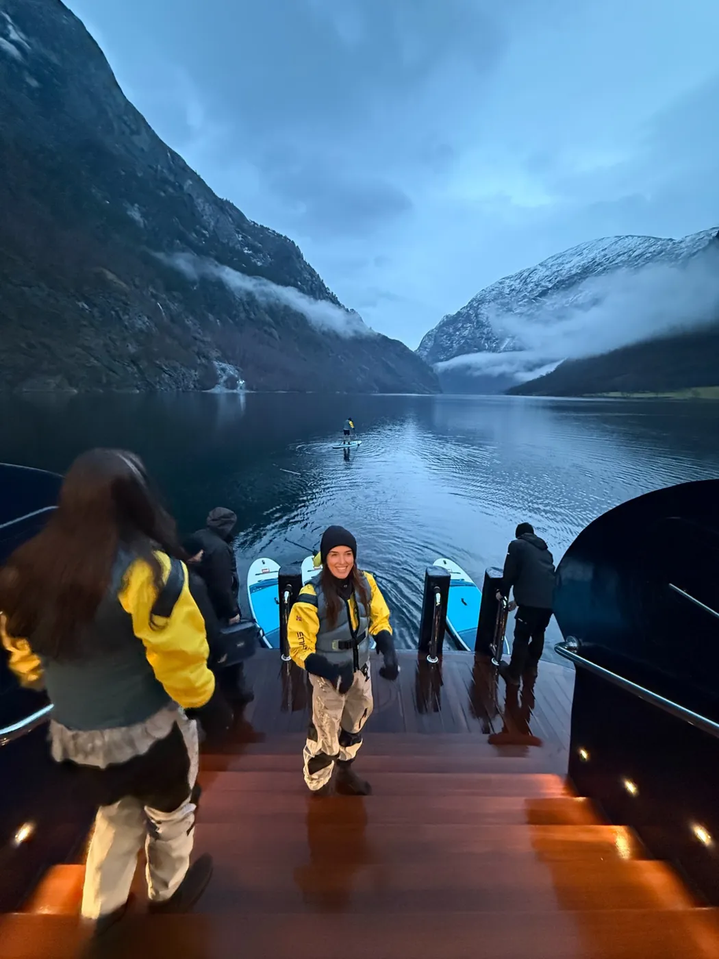 Ellie on ARCTIC's swim platform in a drysuit at dawn, a paddleboarder out on the still fjord behind
